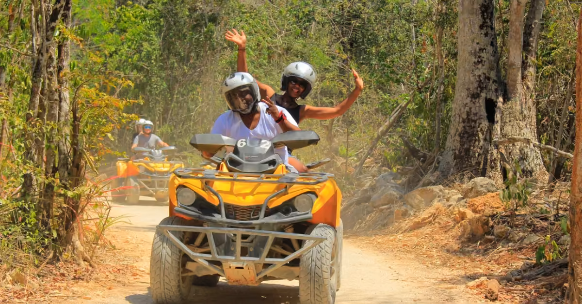 Two helmeted riders smile and wave on a yellow ATV along a dusty forest trail, with another ATV following among the trees.