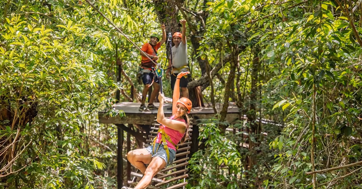 A woman in a pink top and helmet rides a zipline through the Selva Maya forest, smiling and waving at two onlookers behind her.