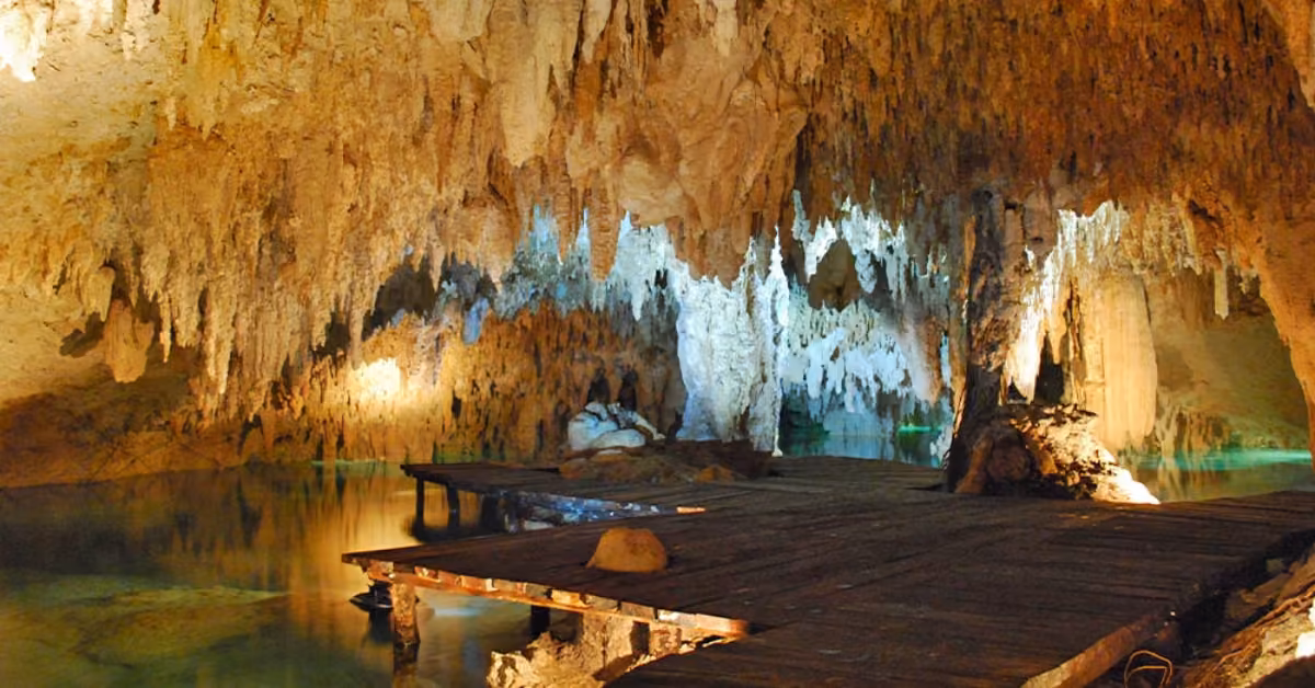 Wooden walkways span clear water inside a cave of striking stalactites and stalagmites, lit by warm and cool lights.