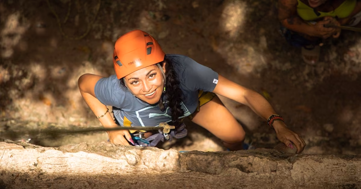 A woman in an orange helmet and climbing gear smiles up at the camera while rock climbing outdoors among sunlit trees.