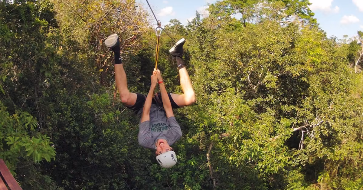 A person in a helmet and harness ziplining upside down above a lush forest, smiling beneath a partly cloudy sky.