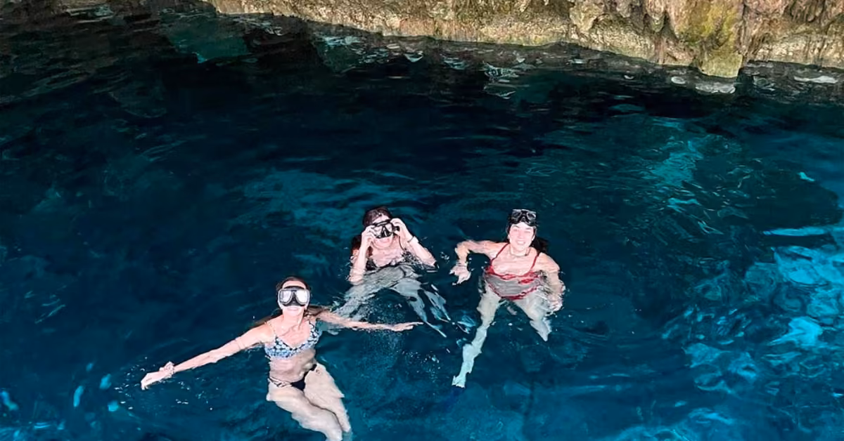 Three friends in swimsuits and snorkels swim in clear blue cave water, smiling up at the camera with rocky walls above.