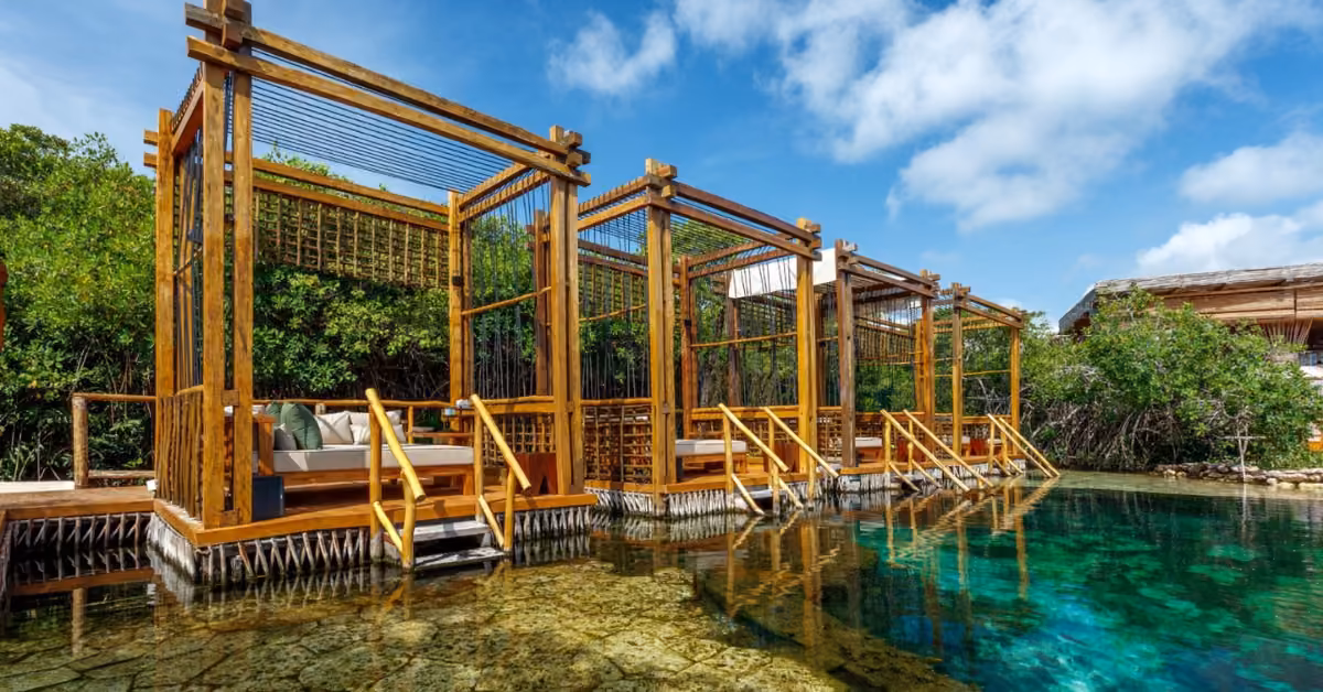Four open wooden cabanas line a clear shallow pool, framed by lush trees and blue sky. Steps from each lead into the water.