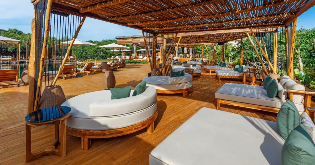 Shibari Cenote Club’s outdoor lounge has cushioned wood seating, pergola shade, greenery, and deck with tables and chairs.