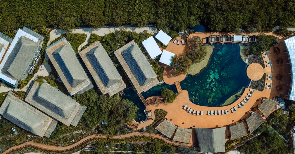 Thatched-roof buildings and a clear pool surrounded by lush greenery, with sun loungers and wooden walkways at Shibari Cenote Club.