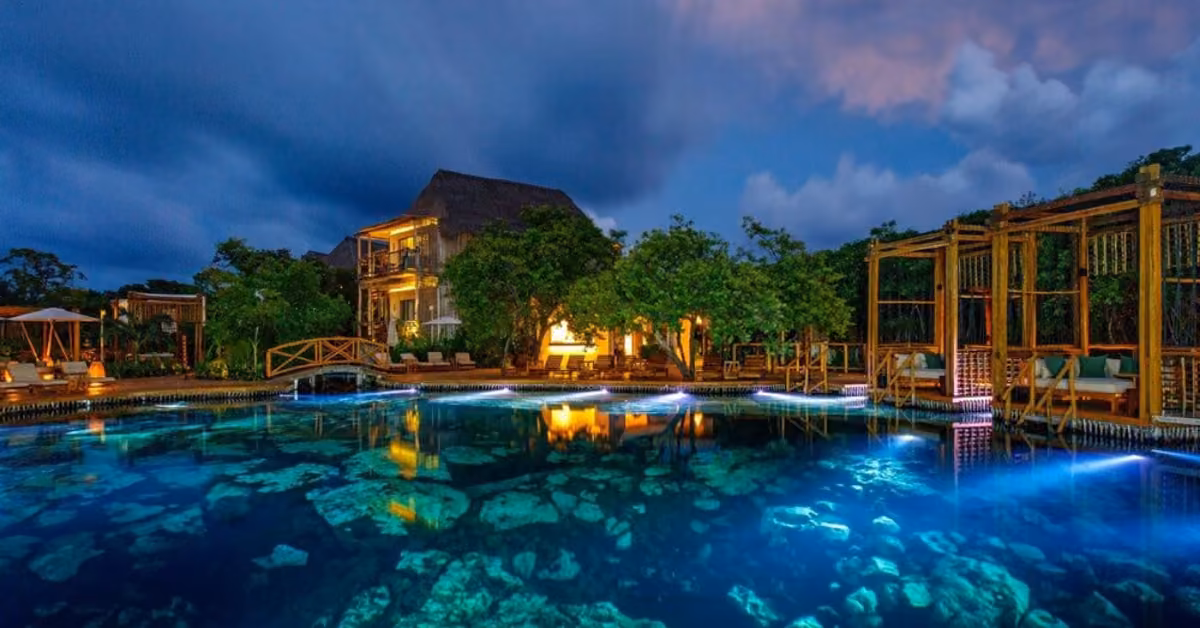 A brightly lit resort at night with a blue pool, reflecting lights, trees, wooden bridges, and elegant cabanas.