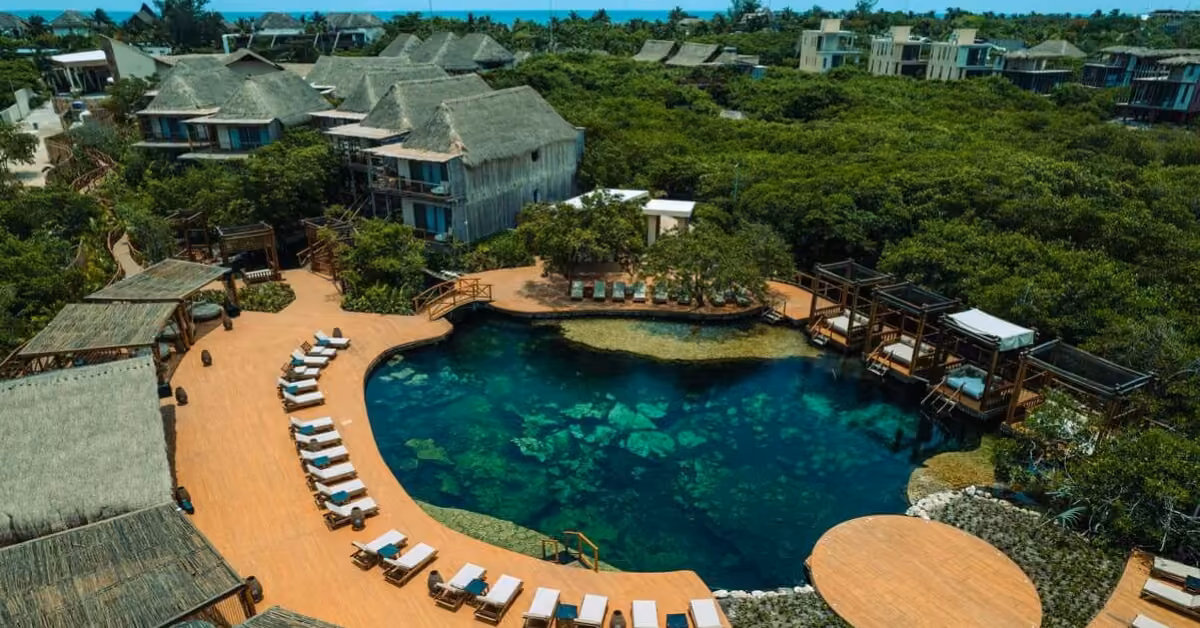 Aerial view of a resort with thatched villas, lush greenery, a blue pool, and lounge chairs lining the wooden deck.