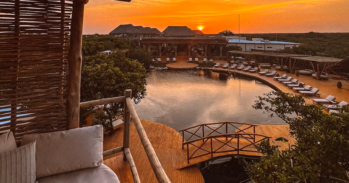 A serene resort pool with a wooden deck and lounge chairs, surrounded by lush greenery at sunset, orange sky reflected in water.