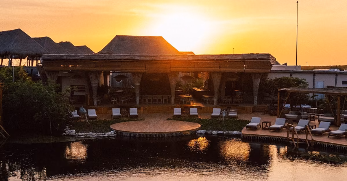 A tranquil resort with thatched roofs and lounge chairs by a reflective pool at sunset, aglow with warm golden light.