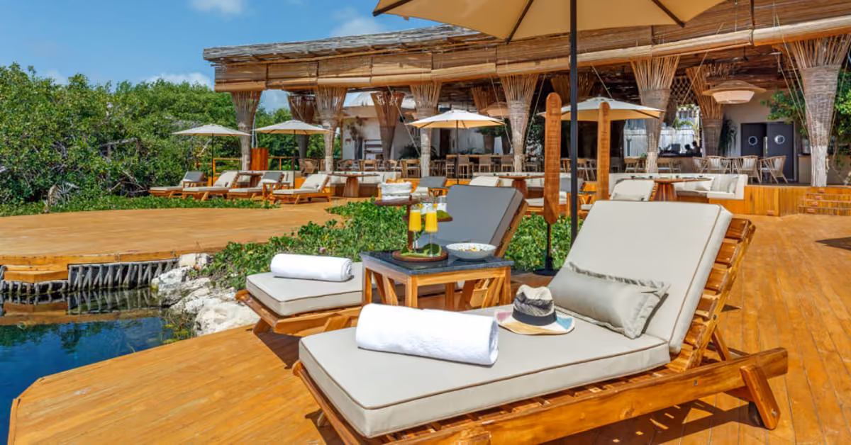 Lounge chairs with cushions, a hat, towels, and drinks on a deck by water near an open-air restaurant amid lush greenery.