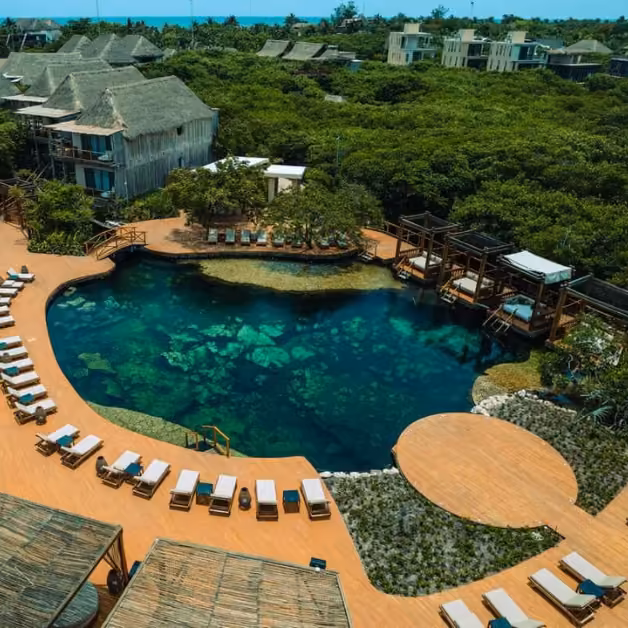 Aerial view of a resort with a lagoon-style pool, lounge chairs, wooden cabanas, lush greenery, and thatched-roof buildings.