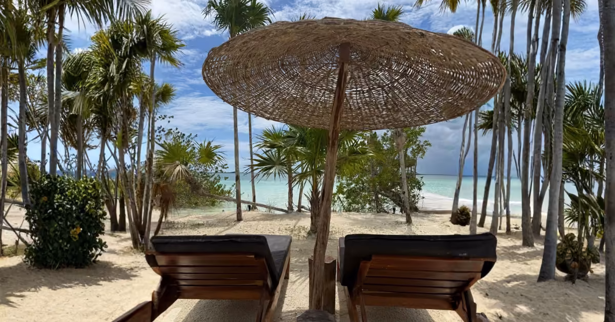 Wooden lounge chairs under a straw umbrella on a sandy beach with palm trees, facing turquoise water and a partly cloudy sky.