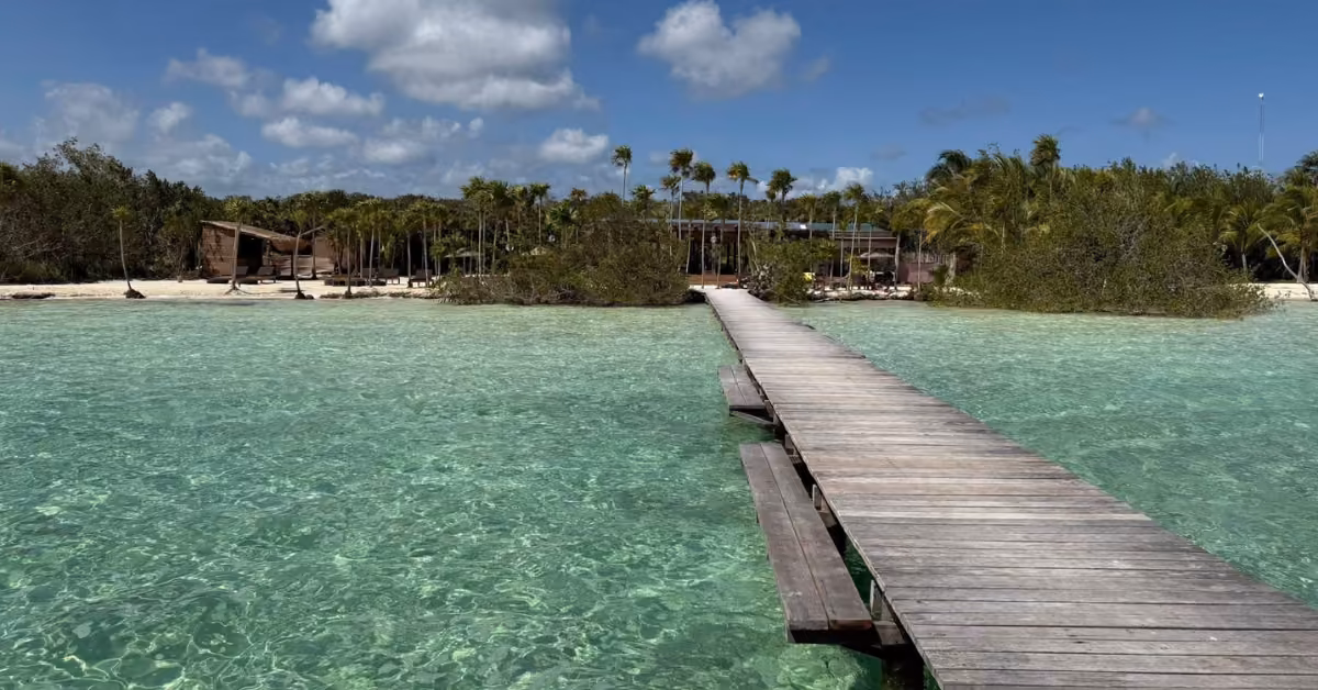 A wooden pier stretches over clear turquoise water to a palm-lined sandy beach beneath a blue sky with scattered clouds.