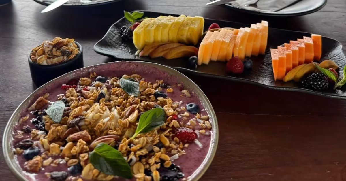 Smoothie bowl with granola, berries, and mint beside plates of papaya, melon, dragon fruit, and a small dish of granola.