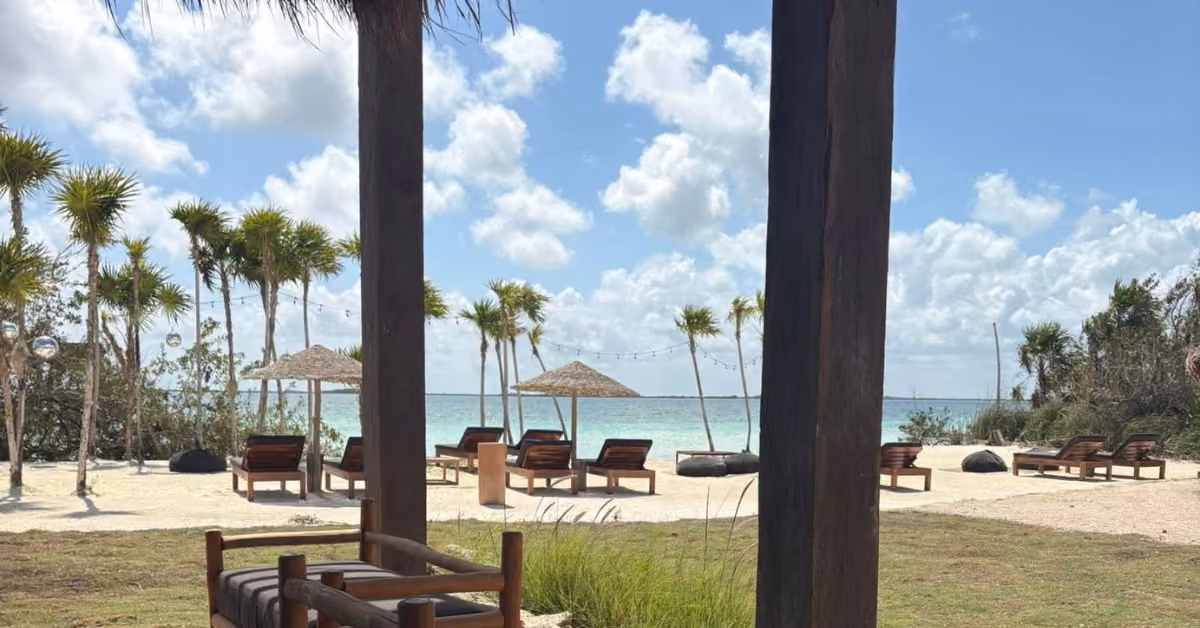 Lounge chairs face the ocean at Sipalitos Lagoon Club, framed by palm trees, straw umbrellas, wooden posts, grass, and sand.