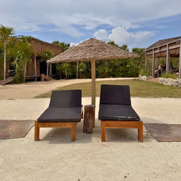 Two wooden lounge chairs with black cushions under a thatched umbrella, surrounded by palms and rustic buildings at Sipalitos Lagoon Club.