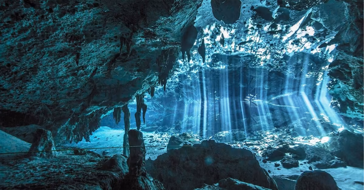 Sunlight filters through clear water in an underwater cave, lighting up rocks and a diver in a serene, blue-tinted scene.