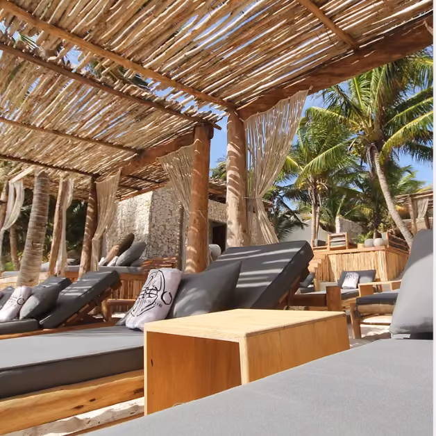 Cushioned lounge chairs and small tables under a rustic pergola, palm trees, and light curtains on a sunny, tropical beach.