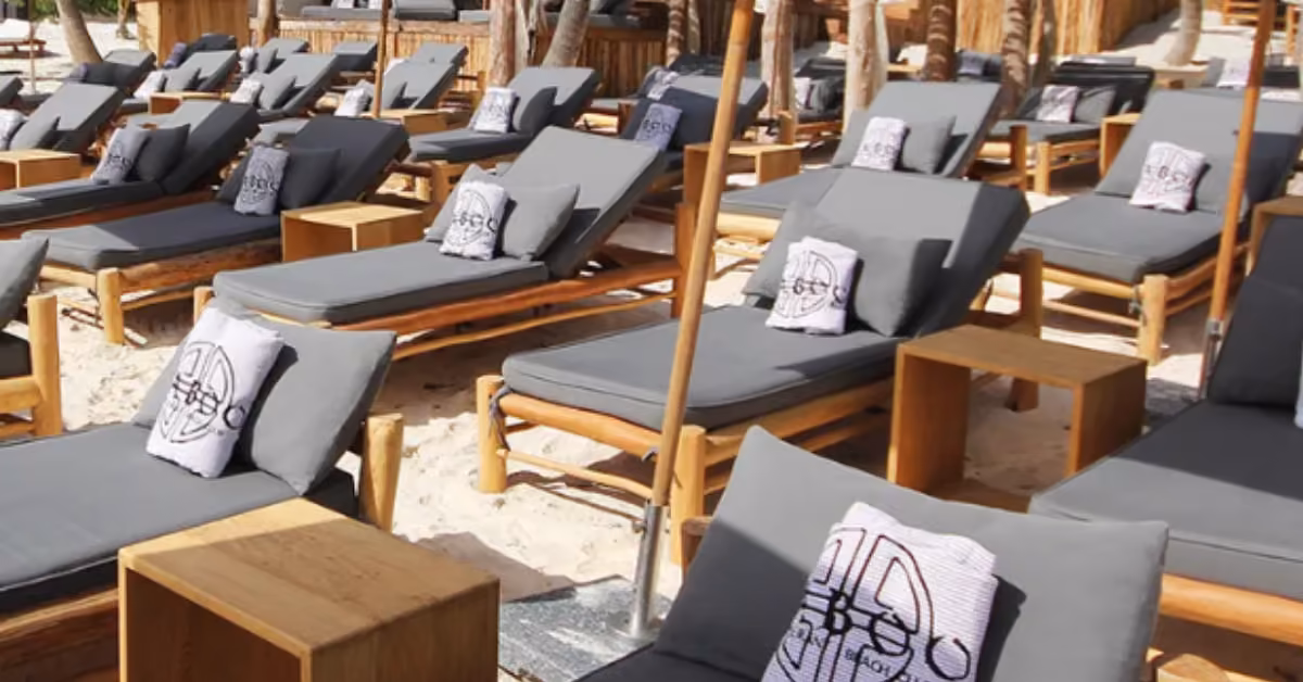 Wooden lounge chairs with gray cushions line sandy ground at Taboo Beach Club, each by a table and large umbrella.