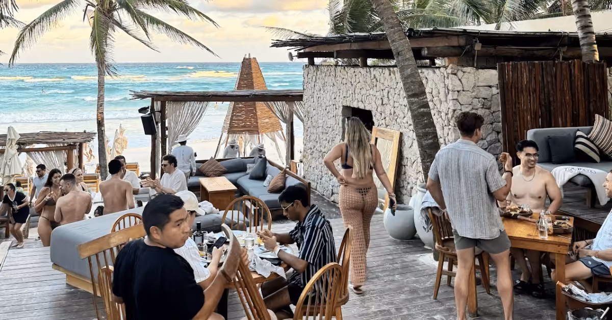 Guests enjoy food and conversation at Taboo Beach Club, a sunny outdoor spot with wooden tables, lounges, palms, and ocean views.