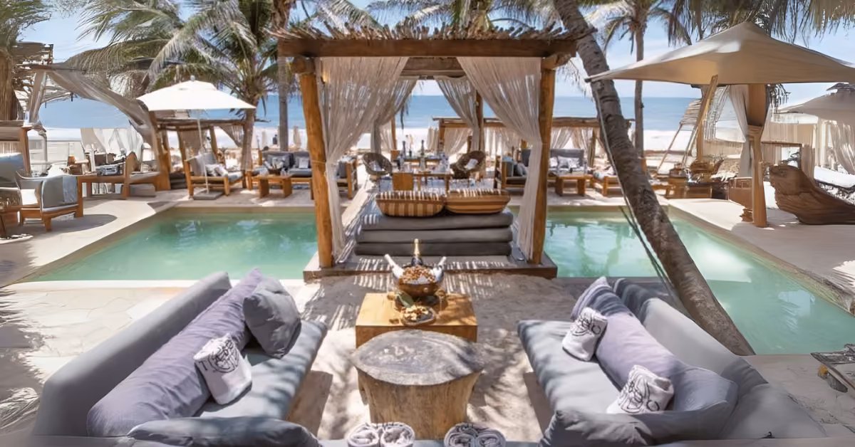 Outdoor lounge with cushioned sofas, pool, wooden tables, and canopy beds under palm trees overlooking a beach and ocean.