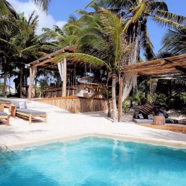 Taboo Beach Club: turquoise pool, cushioned lounge chairs, shaded cabanas, palm trees, and white sand under a sunny sky.