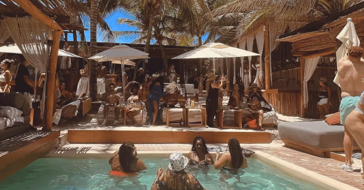 Guests relax and mingle at a sunny pool party with palm trees, a small pool, and shaded cabanas at Taboo Beach Club.