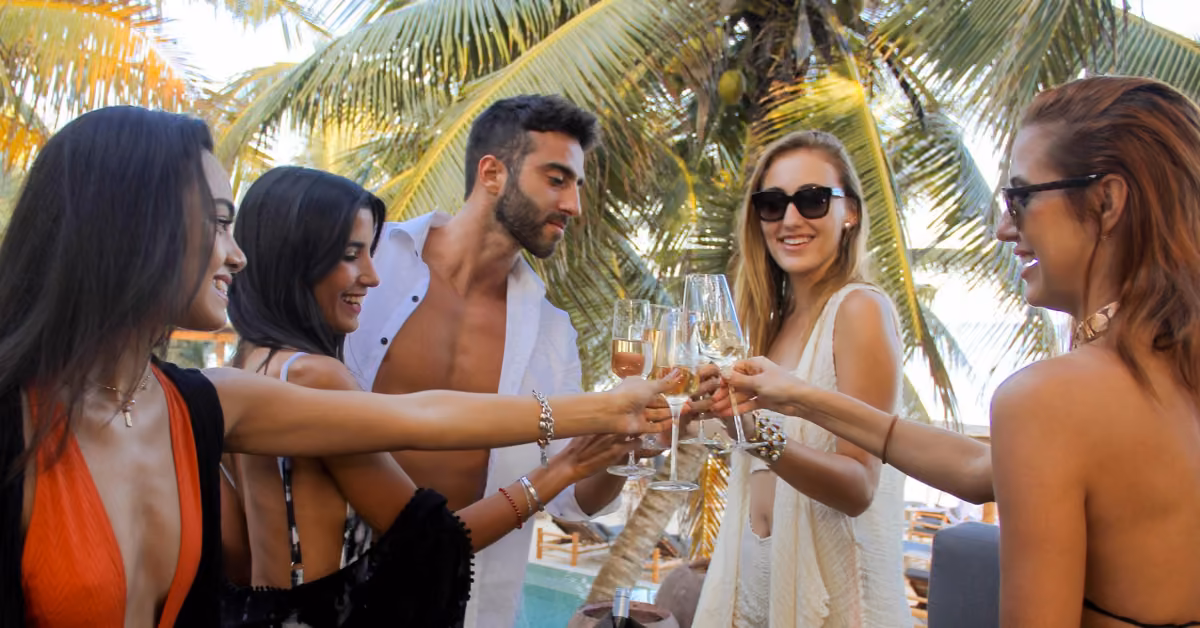 Five people in swimsuits and sunglasses toast with drinks by a pool at Taboo Beach Club, surrounded by palm trees, smiling and enjoying a sunny day outdoors.