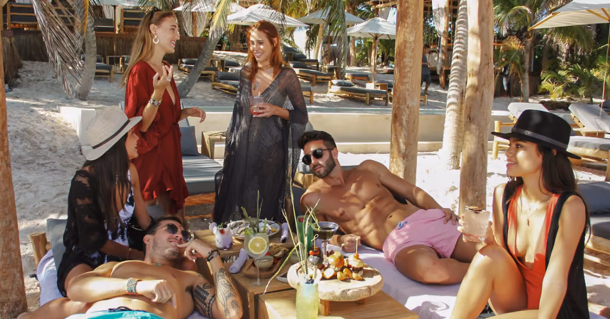Friends lounge at Taboo Beach Club, sharing drinks and snacks by a table under palm trees and wooden shade in the sun.