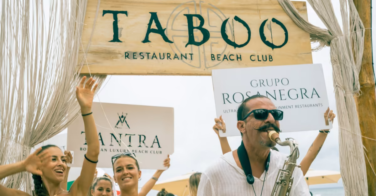 A smiling man plays saxophone at Taboo Beach Club entrance, joined by women holding signs. The group enjoys a lively mood.