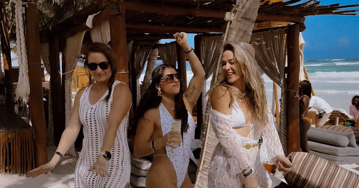 Three women in swimsuits and sunglasses dance and smile on a sandy beach under a cabana, with ocean and palm trees behind.