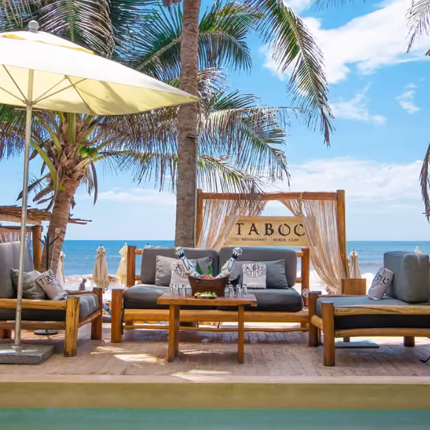 Taboo Beach Club’s outdoor seating has wood furniture and gray cushions, set by palm trees, a pool, and ocean views.