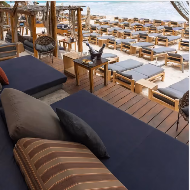 Lounge chairs line the sandy beach at Taboo Beach Club; daybeds and a table with drinks sit on a deck overlooking the view.
