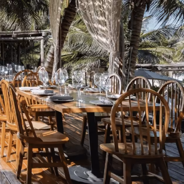 A wooden outdoor table with plates and wine glasses, surrounded by chairs on a deck with palm trees and flowing curtains.
