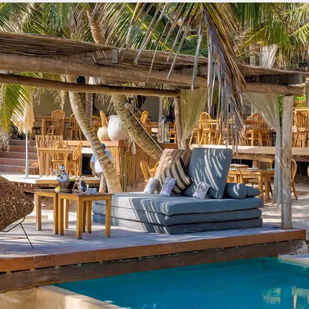 Lounge area with cushioned seats and pillows by a small pool, wooden tables, rustic pergola, and shady palm trees.