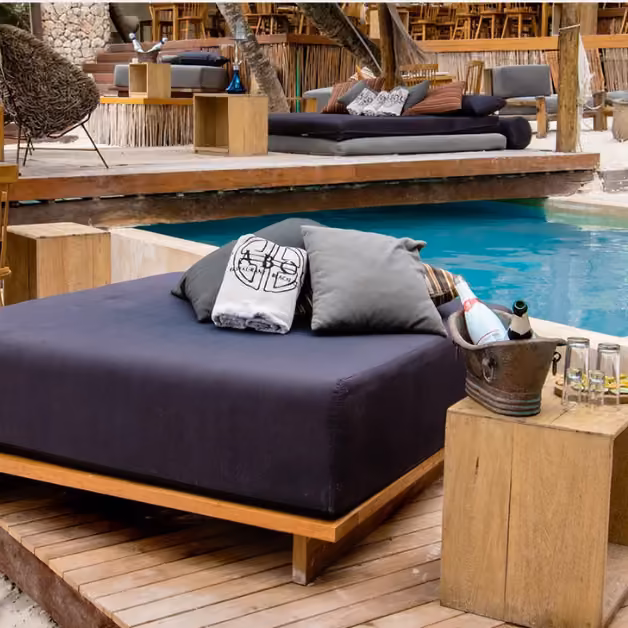 Poolside lounge at Taboo Beach Club with a cushioned daybed, pillows, drinks on a table, and a bar area in the background.