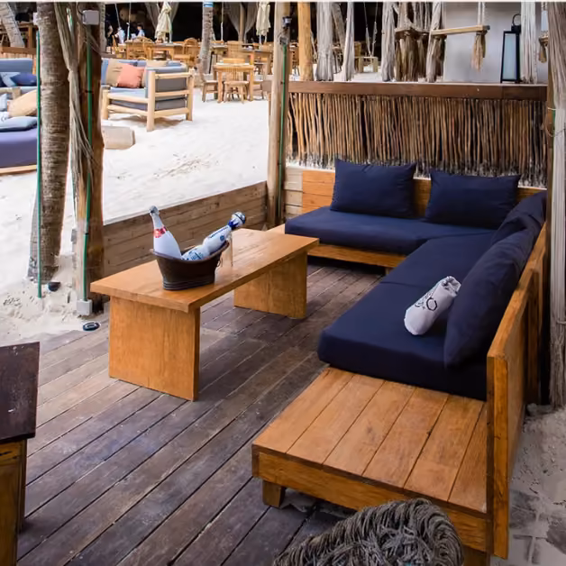 Outdoor lounge at Taboo Beach Club with wooden sofas, blue cushions, a table, wine on ice, and sandy seating in the background.