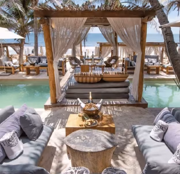 Beachside lounge with cushioned seats, pergola and curtains, shallow pool, palm trees, and ocean view under sunny skies.