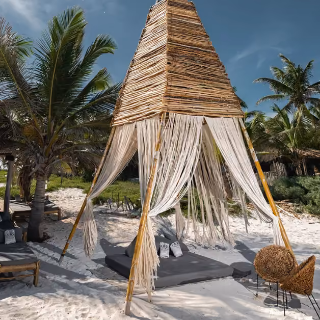 A bamboo cabana with flowing white drapes sits on sandy Taboo Beach, shaded by palms, with lounge seats beneath a clear sky.