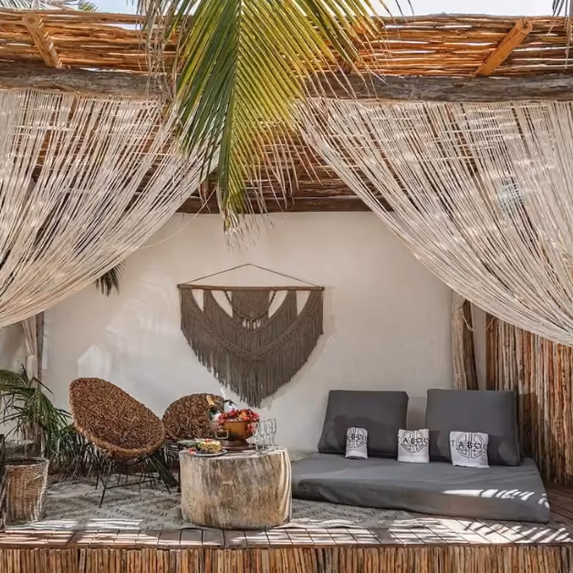 Cozy lounge at Taboo Beach Club with woven chairs, wood table, cushions, pillows, macramé art, and draped pergola.
