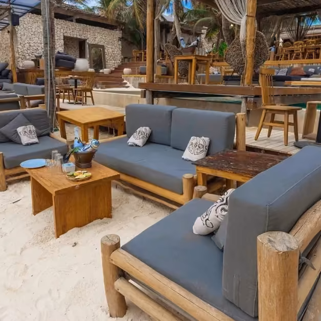 Beach lounge with wooden furniture, gray sofas, throw pillows, small tables on sand, palm trees, and open-air dining.