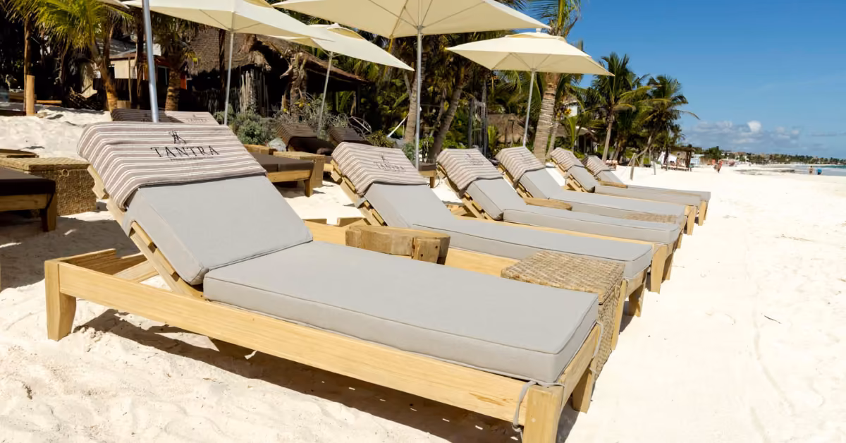 Wooden lounge chairs with gray cushions and striped pillows sit on sand under white umbrellas, palms and blue sky behind.
