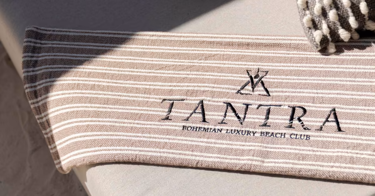 A beige and white striped towel with black embroidered text, laid on a light surface beside a textured cushion.