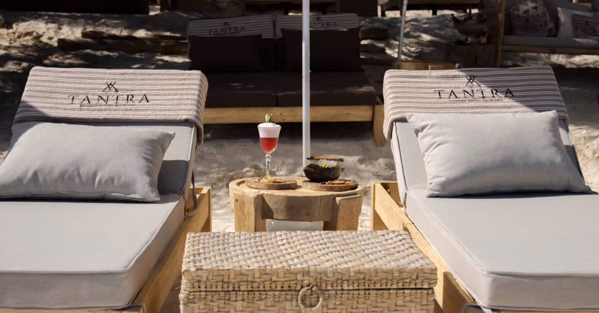 Two cushioned lounge chairs with branded towels face a table with a pink drink and snacks on a sandy beach, more chairs behind.