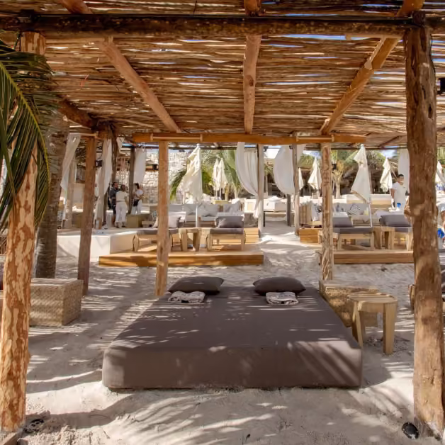 Shaded beach lounge at Tantra Beach Club with wooden pergolas, cushioned beds, palm trees, and flowing white drapes.