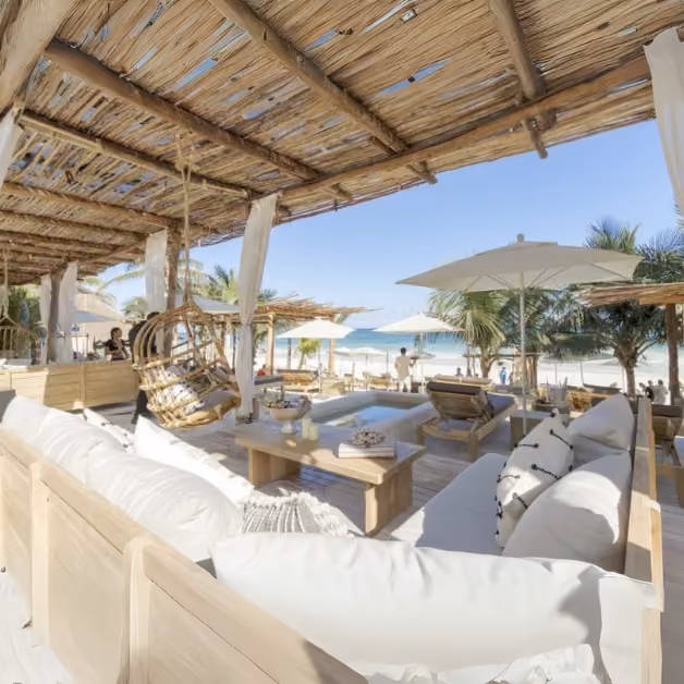 Tantra Beach Club lounge with white sofas, wooden tables, and palm trees. Guests relax by the sea under umbrellas and a bamboo roof.