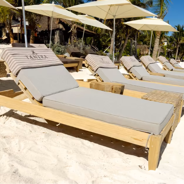 Cushioned lounge chairs with striped towels sit on the sand under white umbrellas, backed by palms and lush greenery.