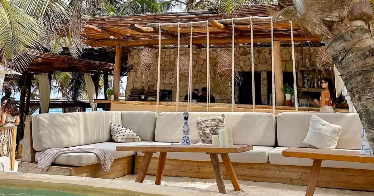 Tantra Beach Club’s outdoor lounge offers cushioned seats, wooden tables, and pillows under palm trees by a rustic stone bar.
