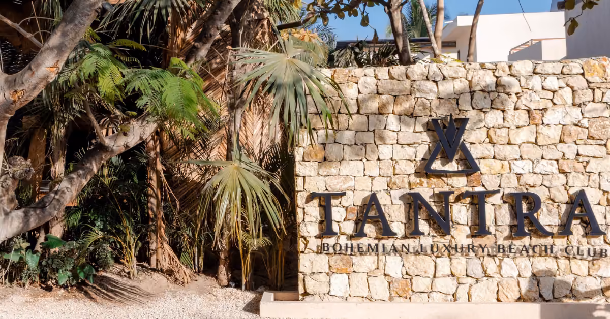A stone wall sign for Tantra Beach Club with palm trees and greenery, welcoming guests in a sunny outdoor setting.