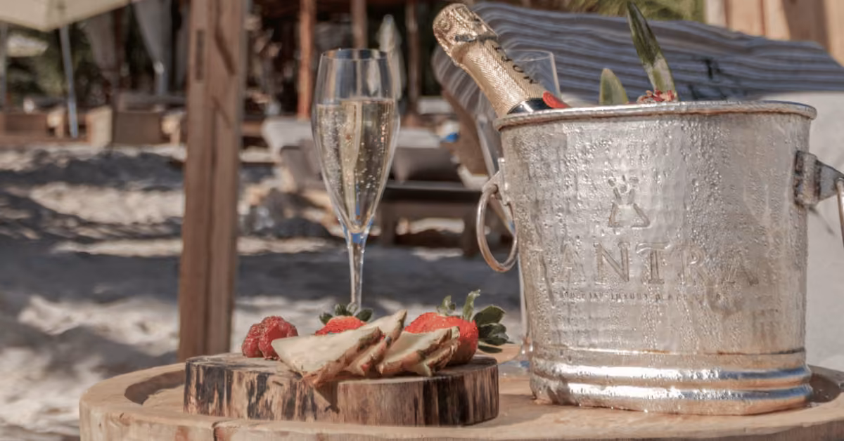 Champagne on ice, a filled flute, and strawberries on a wooden board sit atop a rustic table by the sea at Tantra Beach Club.